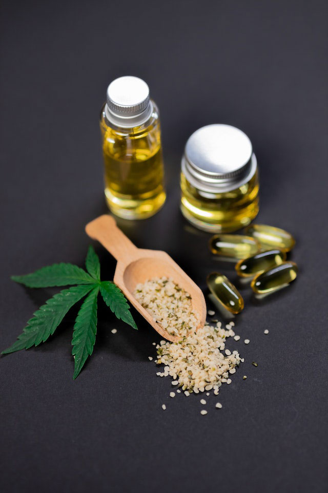 A display of two bottles of CBD tincture, CBD capsules, a brown wooden scoop with hemp seeds, and a cannabis leaf on a charcoal grey background