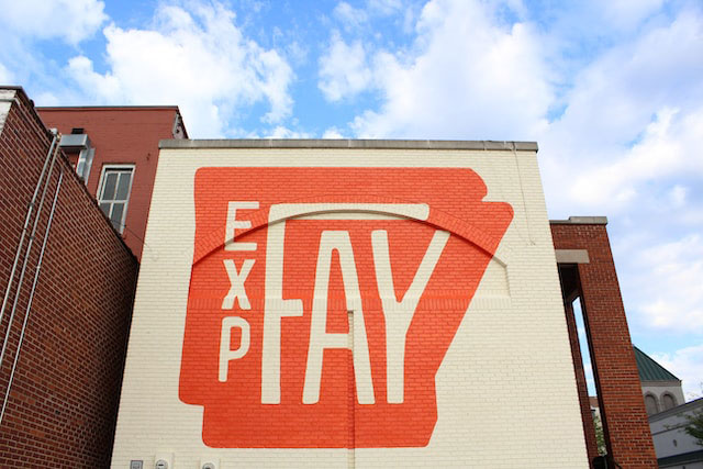 Large mural on the side of brick building in Fayetteville that reads "EXP FAY" in cream with orange background