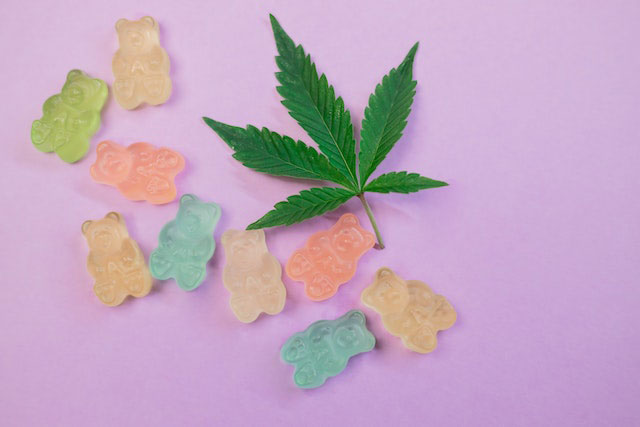 Pastel-colored gummy bears next to a marijuana leaf all lying on a pink/lavender background