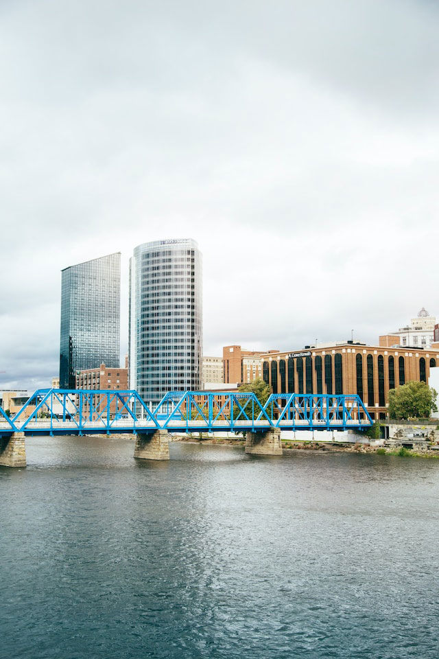 Blue steel frame bridge over a body of water with office buildings behind it in Grand Rapids, MI