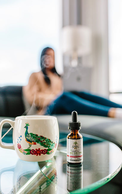 A bottle of CBD oil next to a mug with a peacock on it and steam coming from the top. A blurry image of a person is in the background