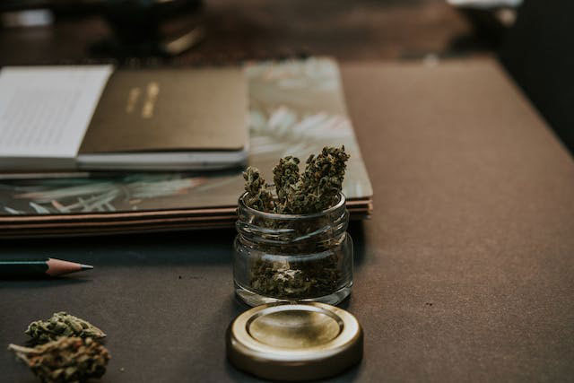 Cannabis in a glass jar on a brown desk with a pad and pen