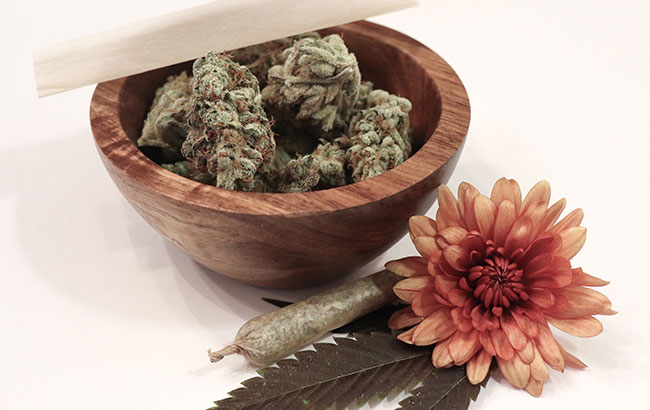 A small wooden bowl full of weed buds. a rolled joint and a lovely orange flower.