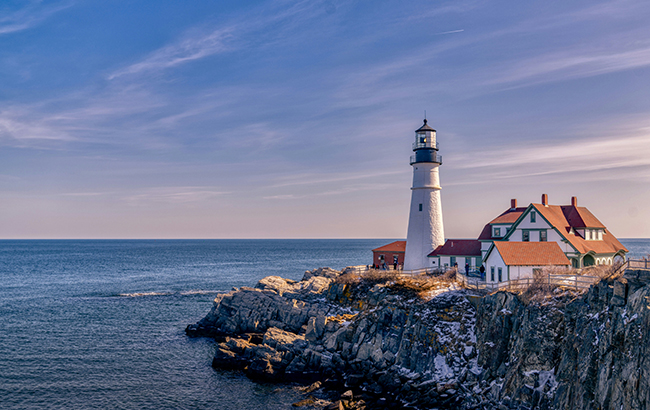 a beautiful picture of the Maine coast. a dark blue ocean against a light pink sunset and a white lighthouse on the jagged cliffs.