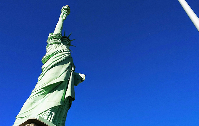 statue of Liberty next to American flag blowing against blue sky